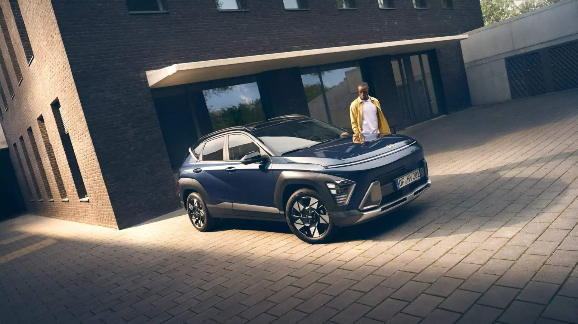Mörkblå Hyundai KONA Hybrid parkerad på en stensatt gård, en man lutar sig mot motorhuven