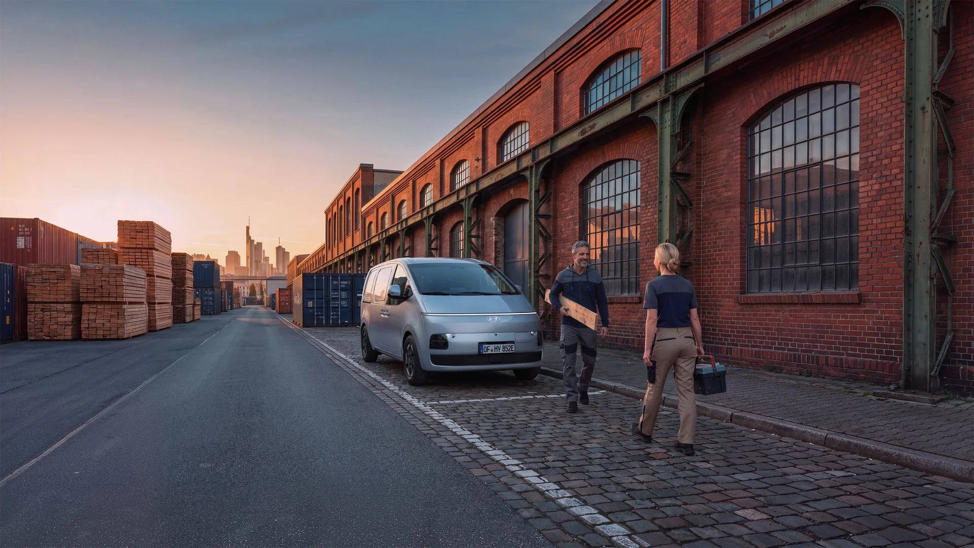 Hyundai STARIA Cargo parkerad utanför en tegelbyggnad, med två personer och arbetsutrustning bredvid bilen.