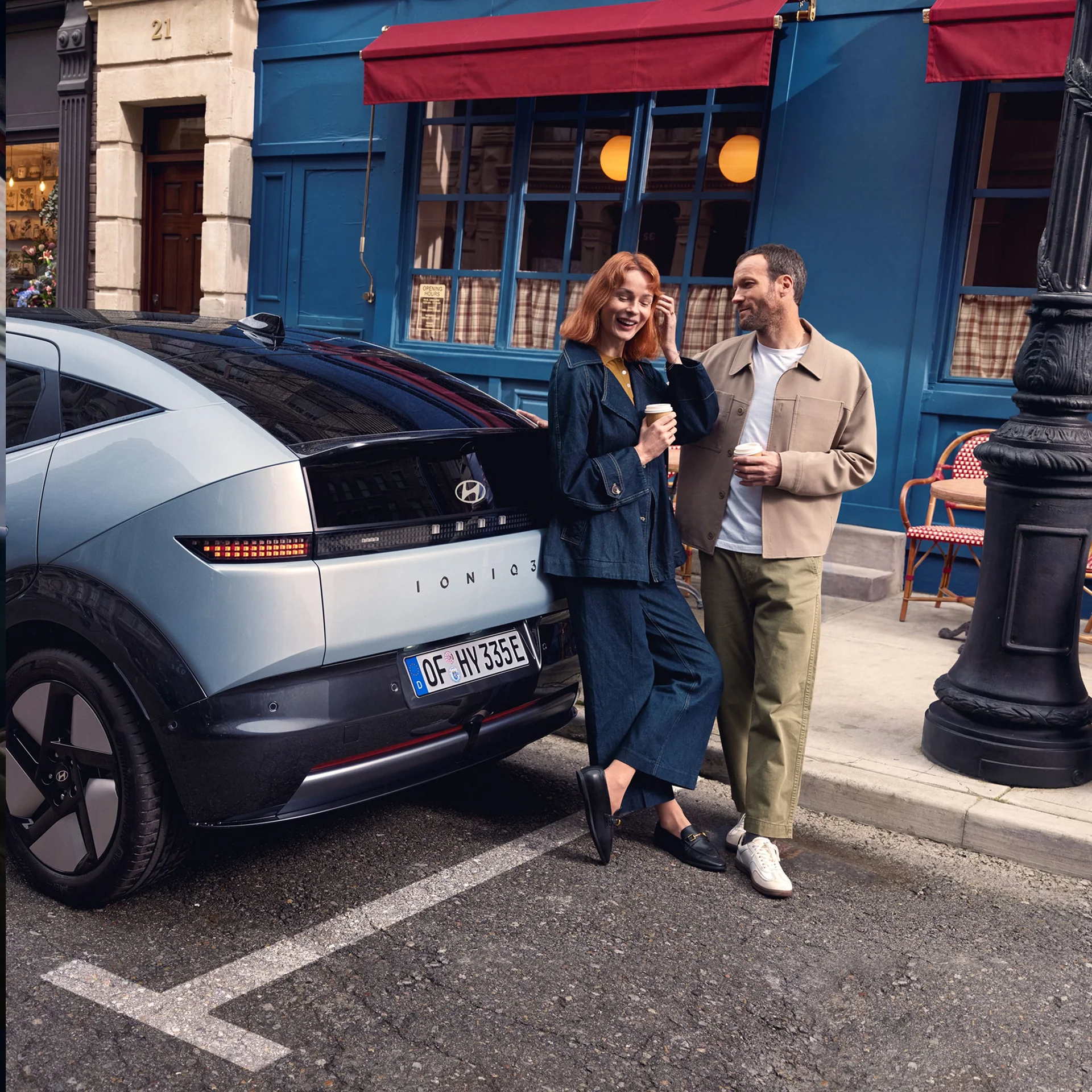 Ett par håller i kaffemuggar bredvid en Hyundai IONIQ 3 på en färgglad gata