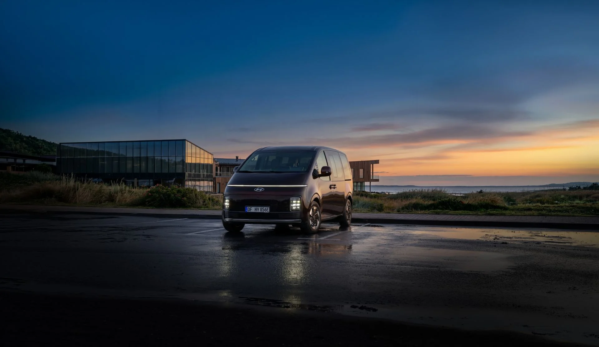 Hyundai STARIA Electric sedd framifrån i skymning, parkerad framför en modern byggnad vid havet